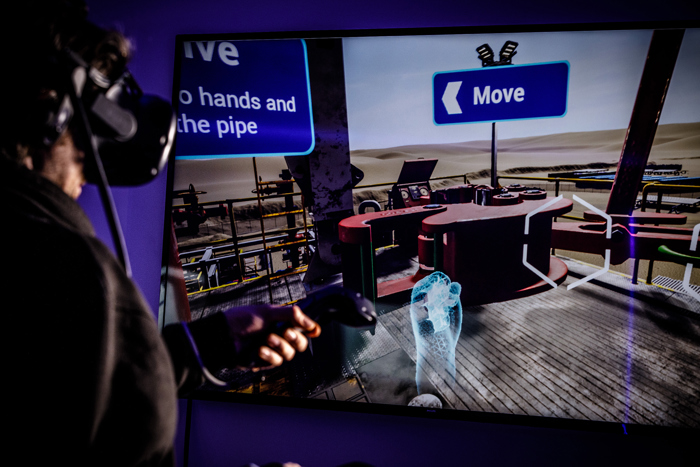 Person using a virtual reality headset and controllers, interacting with an industrial simulation on a screen with instructions to move and use hands and pipe.