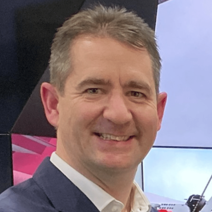 A man with short brown hair wearing a navy blazer and white shirt, smiling in front of a backdrop featuring modern design elements and technological equipment.