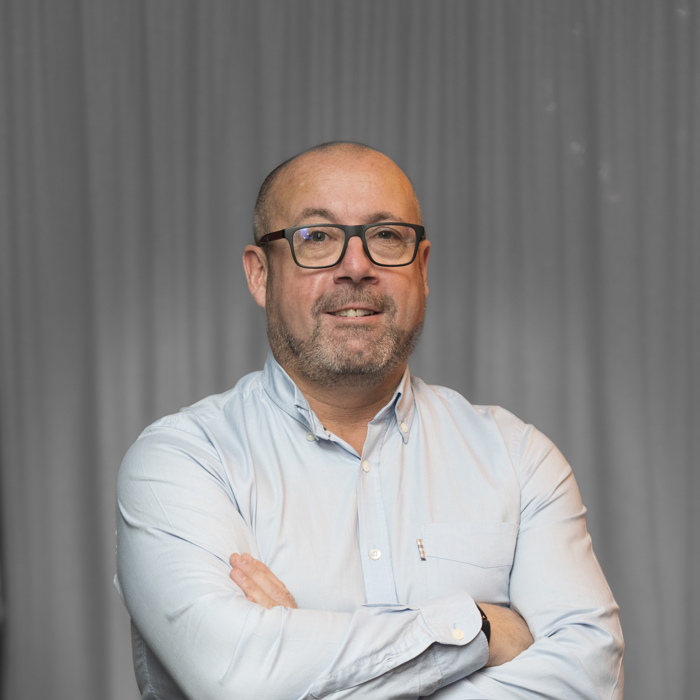 A bald man with glasses and a beard, wearing a light blue button-up shirt, stands with his arms crossed in front of a gray curtain.