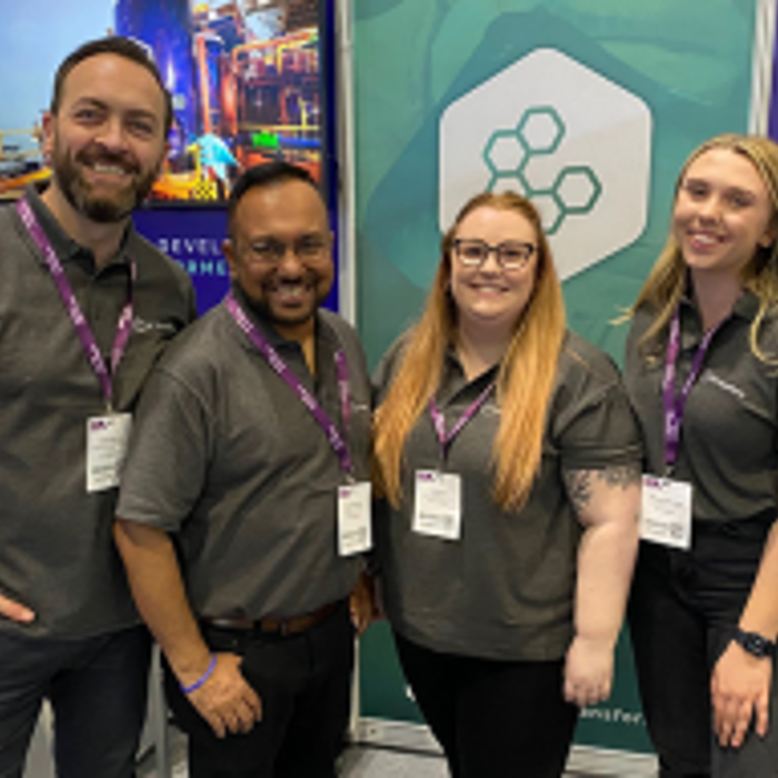 Four people wearing matching shirts stand together, smiling, in front of a display booth featuring promotional banners at an event or conference.