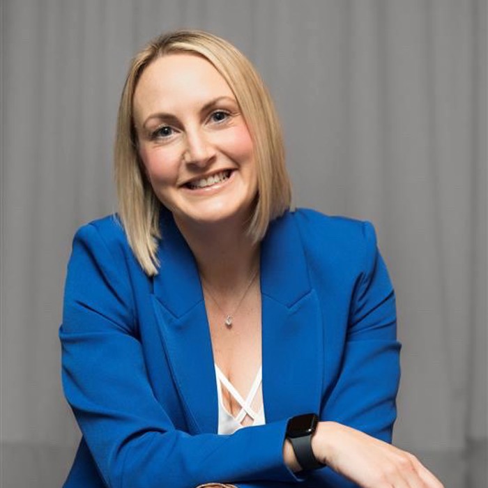A person wearing a blue suit and a smartwatch, smiling while seated with arms resting on knees, and a gray curtain in the background.