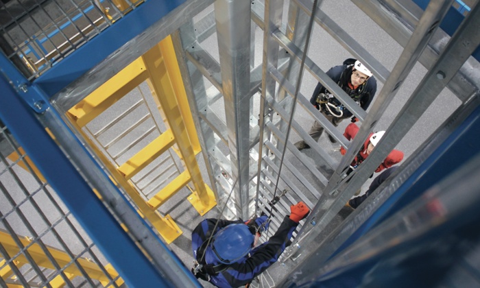 Workers equipped with safety gear are climbing a metal structure. One worker is ascending while others follow closely below, surrounded by metal beams and a yellow ladder frame, following protocols from 3t Training Services.
