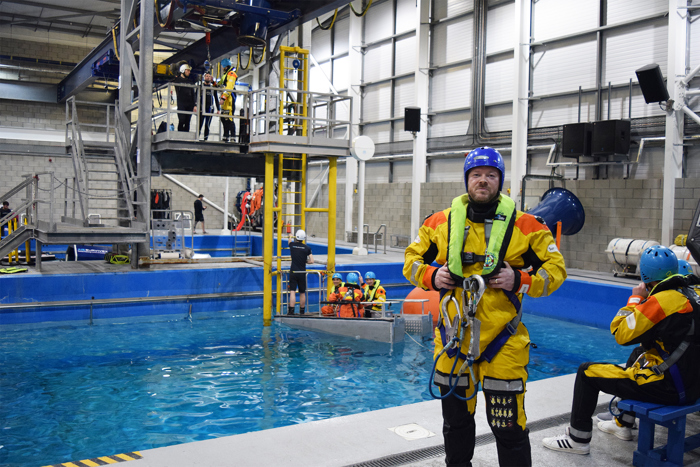 A person in a protective marine suit stands near a pool in an indoor training facility, conducting essential training exercises. Others, dressed similarly, are seen participating actively in and around the pool area.