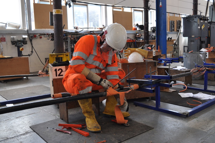 A worker in orange protective gear and a white helmet repairs equipment in a workshop, surrounded by tools and supplies.