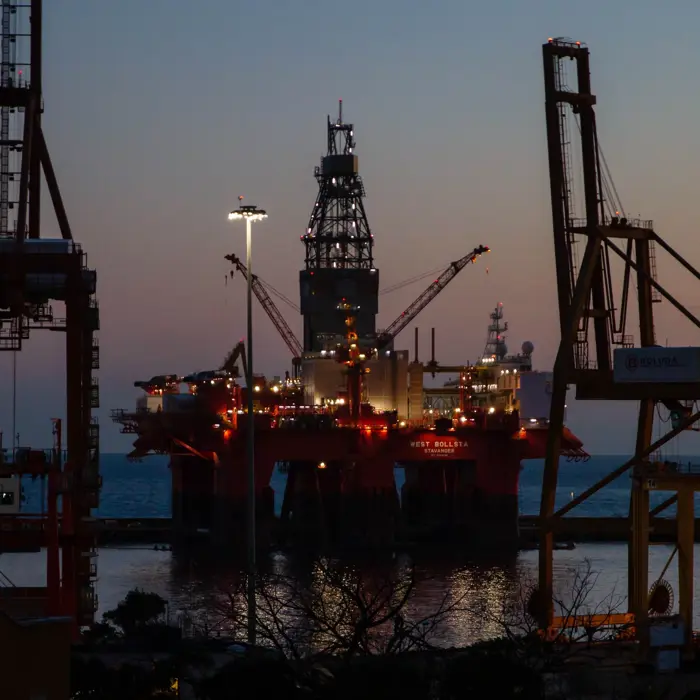 An offshore oil rig lit up at dusk, surrounded by the silhouettes of large cranes and industrial structures, with the sea in the background.
