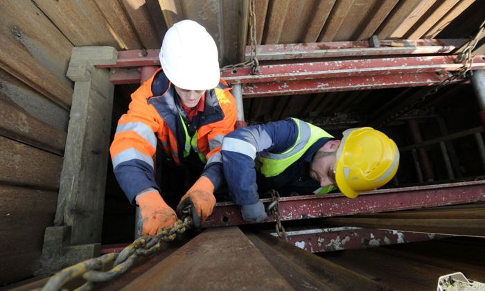 Two construction workers in safety gear are handling chains inside a narrow trench, surrounded by metal reinforcements.