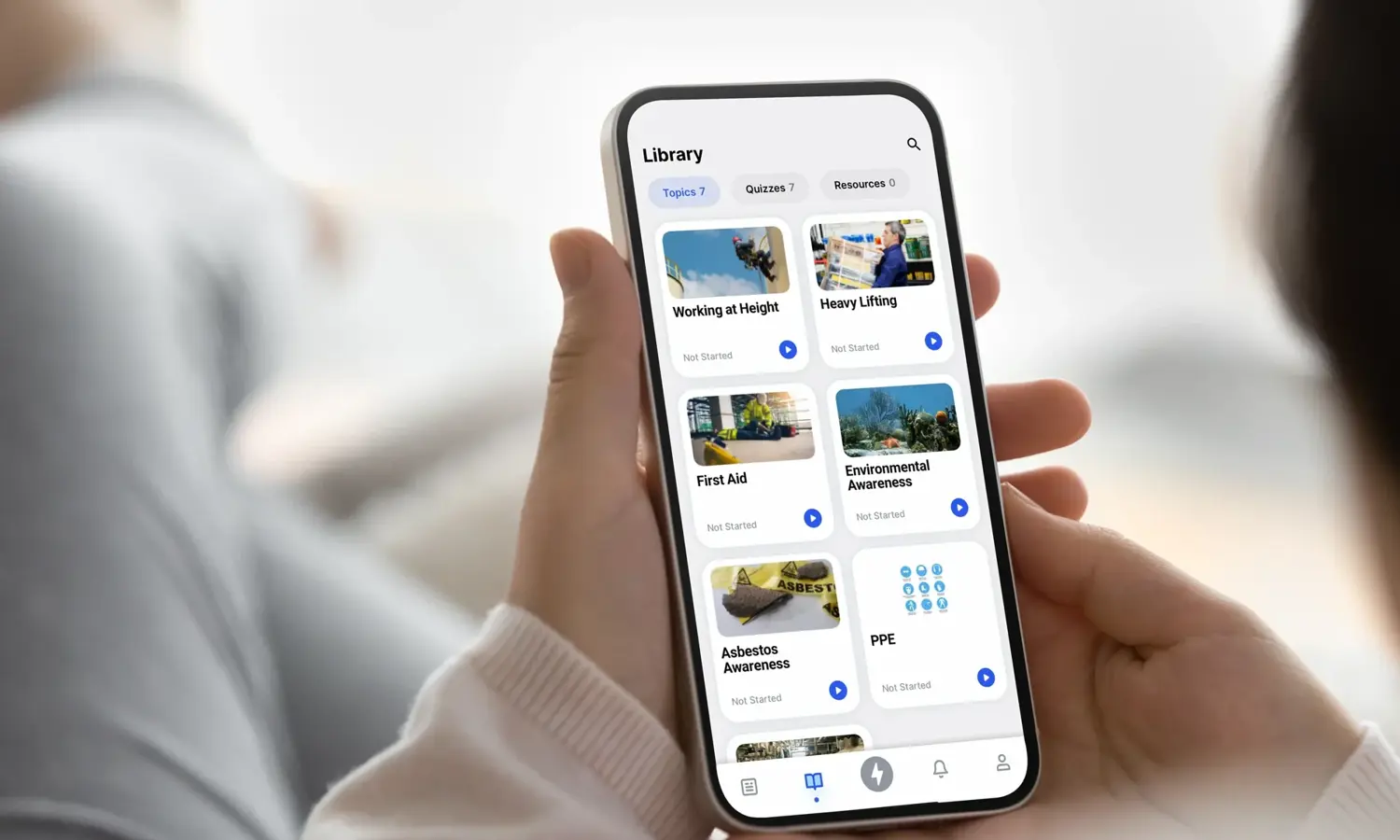 Person using a smartphone app to browse a library of courses including "Working at Height," "First Aid," "Asbestos Awareness," among others, with options categorized under various topics and sectors.