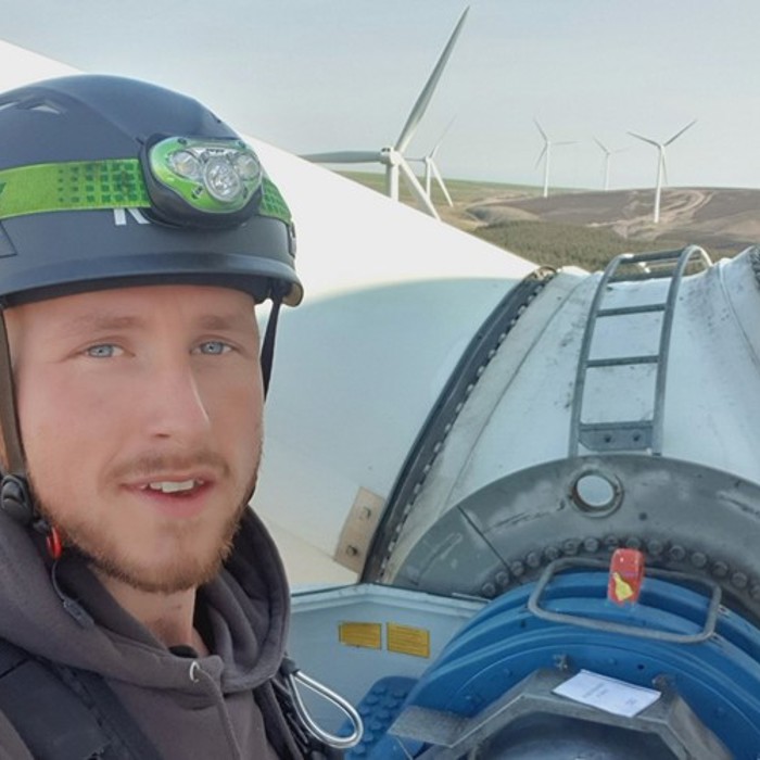 A person wearing a helmet and safety gear stands in front of a wind turbine, with multiple wind turbines visible in the background.