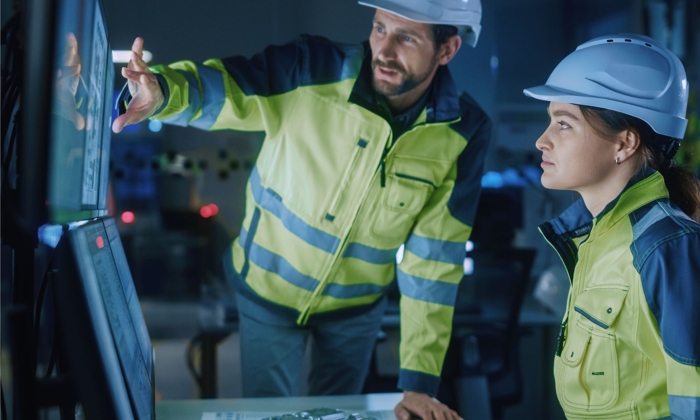 Two engineers wearing hard hats examine data on computer screens in a control room, with one pointing at the screen and the other attentively observing.