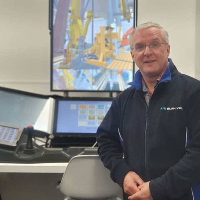 A man wearing glasses and a navy jacket stands in front of a control station with multiple screens displaying industrial equipment.