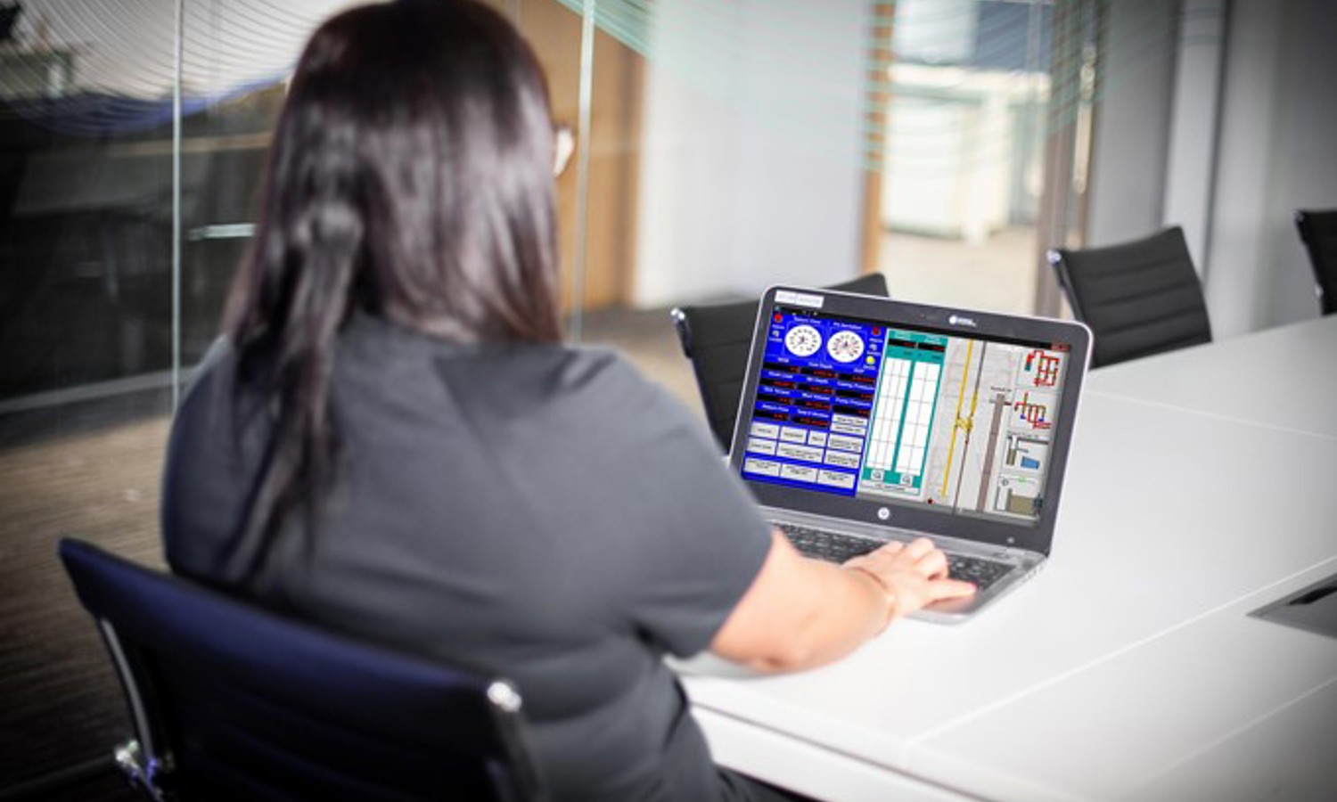 A person with long dark hair sits at a conference table, working on a laptop with a technical interface displayed on the screen.