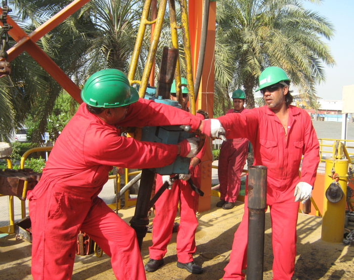 Workers in red coveralls and green helmets operate machinery on an oil rig, surrounded by palm trees.