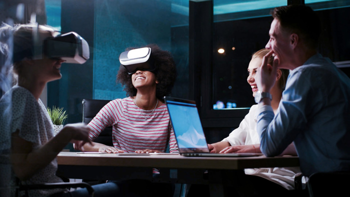 Four people sit at a table in a dimly lit room, two wearing virtual reality headsets, laughing and appearing to enjoy the experience. A laptop is open on the table.