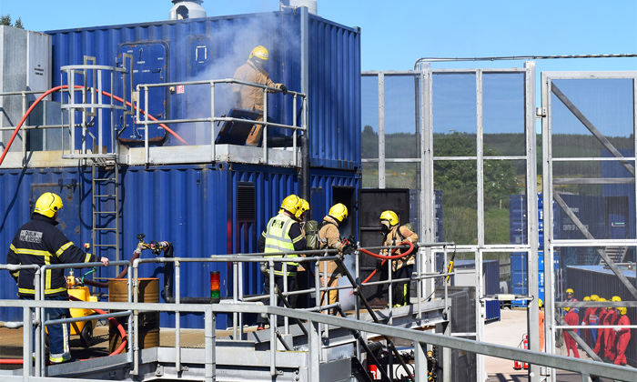 Firefighters in yellow and black uniforms conduct a training exercise, using hoses and equipment on a multi-level blue structure. Smoke is visible from the upper levels.