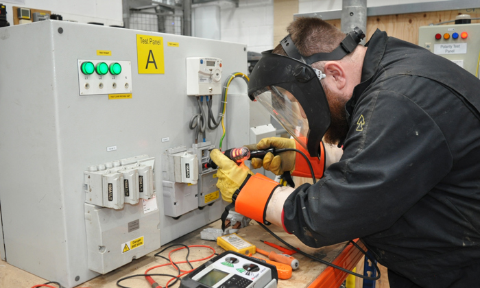 A person wearing protective gear engages in training, working on an electrical test panel with tools and meters spread across a wooden table.