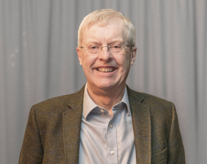 An older man with glasses and a gray jacket smiles confidently, possibly reflecting on years of training, in front of a gray curtain.