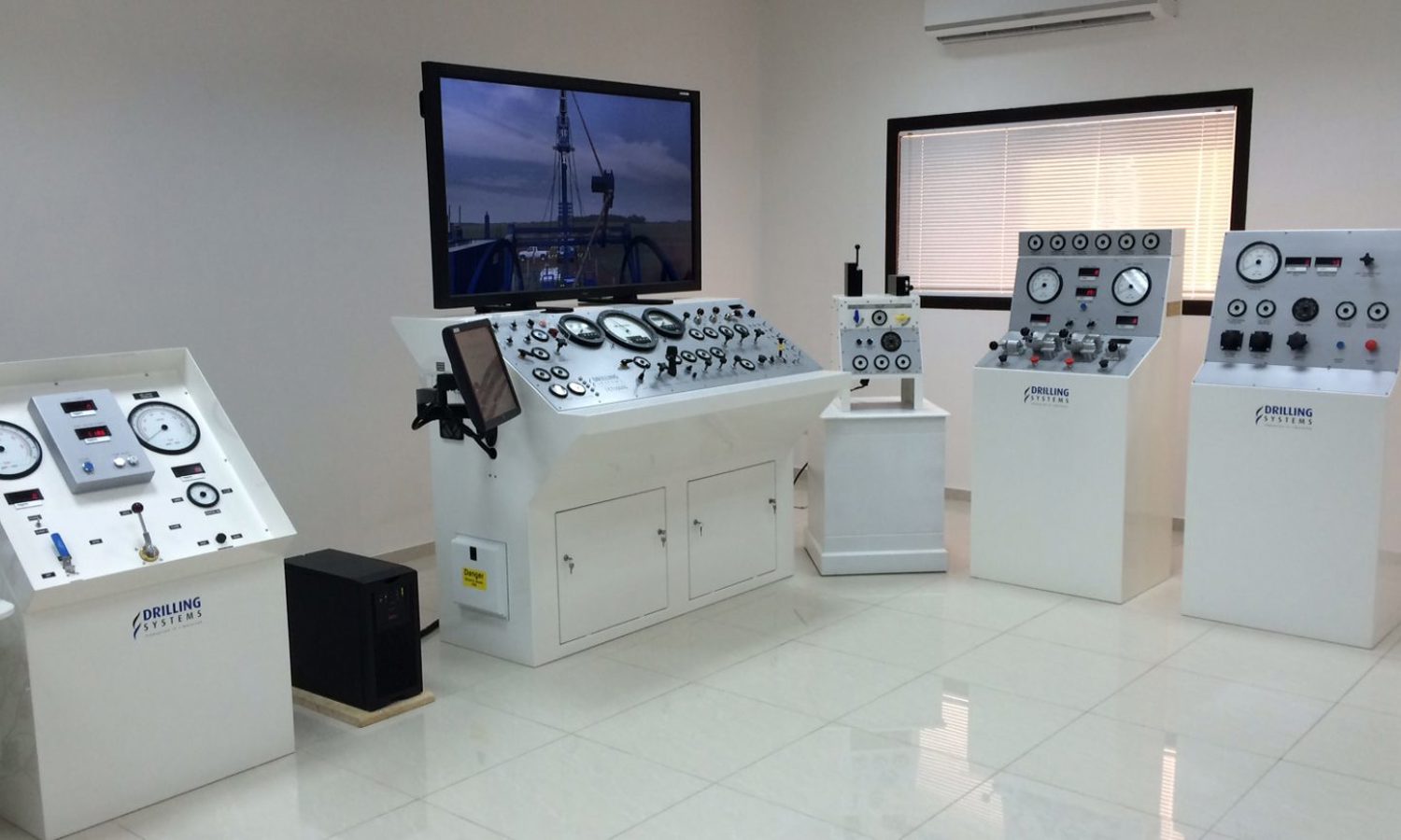 A room with multiple control panels, gauges, and a large screen on the wall. The equipment is arranged on white stands and cabinets, and the floor is tiled.