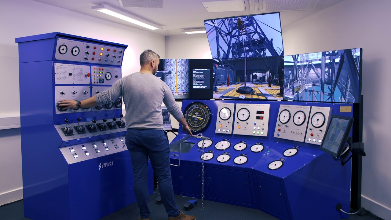 A person operates a blue industrial control panel with multiple screens and gauges, likely in a simulation or control room environment.