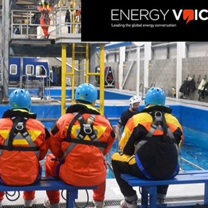Three people in protective gear and helmets seated on a bench near a training pool in an industrial facility, with "ENERGY VOICE" logo and tagline in the top right corner.