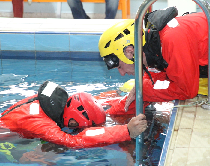 Two rescue workers in red suits and helmets perform a pool training exercise, with one standing in the water holding the pool edge and the other leaning over.