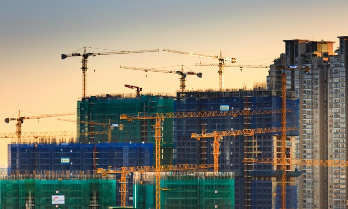 High-rise buildings under construction with multiple cranes and green netting, set against a sunset sky.