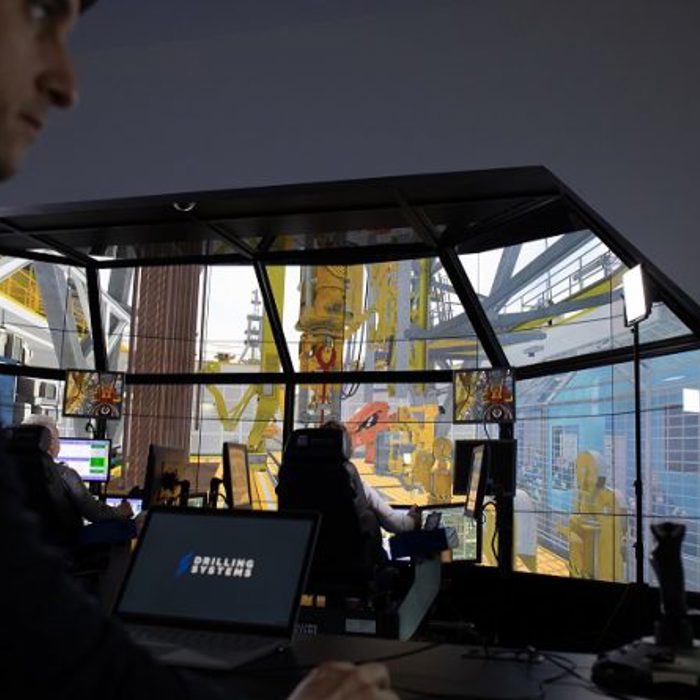 A person operates a computer with DrillSIM software as others use a drilling simulator featuring multiple screens and industrial visualizations.