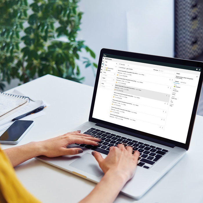 Person typing on a laptop at a desk with an open email inbox on the screen, next to a notebook, pen, smartphone, and a glass of water. A plant is in the background.