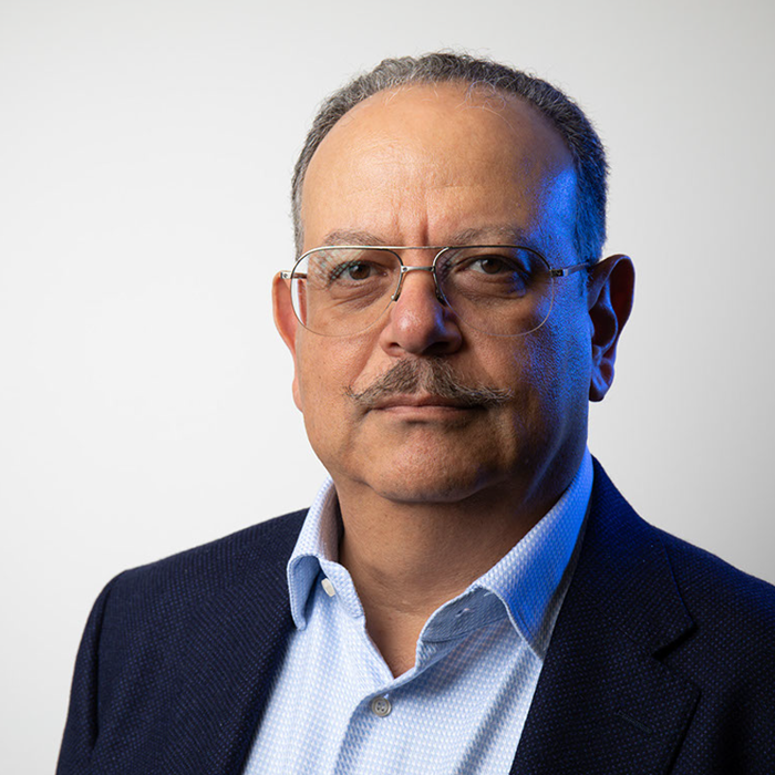 A middle-aged man with short hair, wearing glasses, a light blue shirt, and a dark blazer, is looking directly into the camera. The background is plain and light-colored.