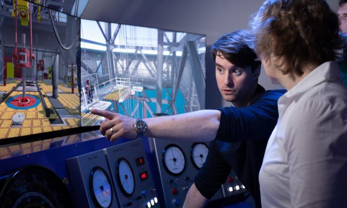 Person points at a screen displaying industrial controls and machinery simulations while explaining to another individual in front of control panels with analog gauges.