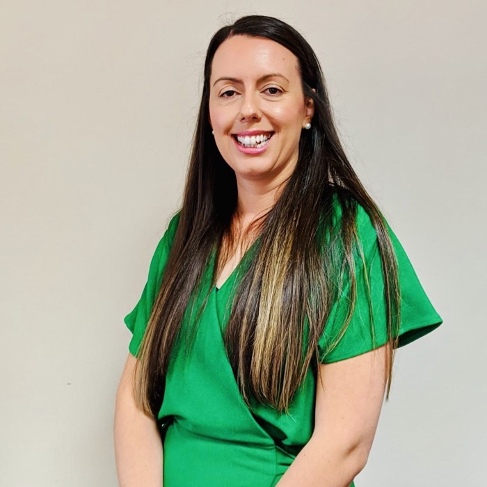 A woman with long brown hair wearing a green dress stands against a plain background, smiling at the camera.