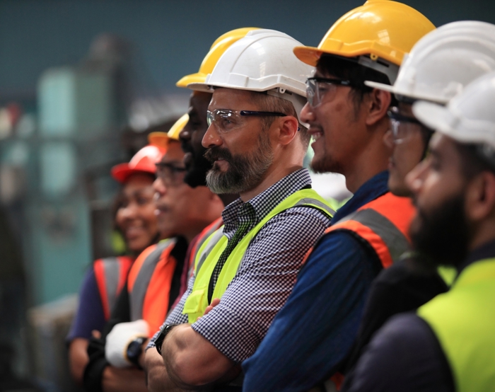 A group of workers wearing safety helmets and vests stands together, focusing intently on a training exercise just out of view.