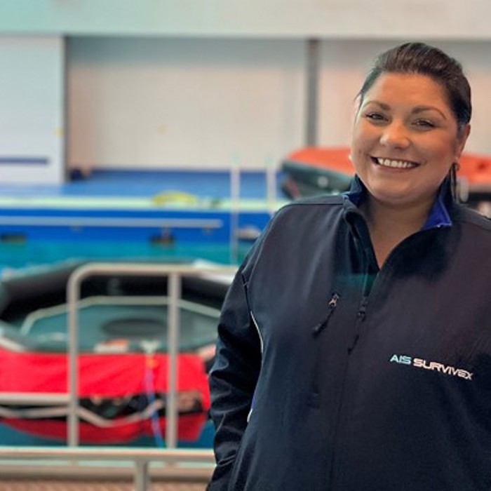 A person in a navy blue jacket stands smiling in front of a pool equipped with lifeboats and safety equipment.