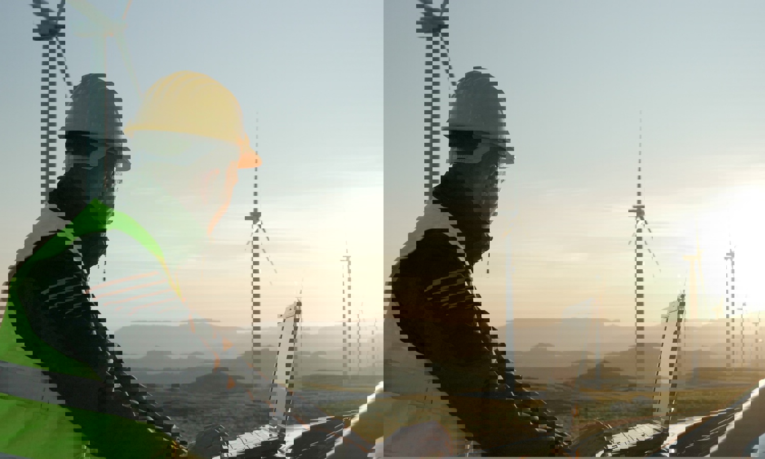 Person in a hard hat and safety vest using a laptop outdoors near wind turbines, with a scenic hilly landscape in the background during sunset.