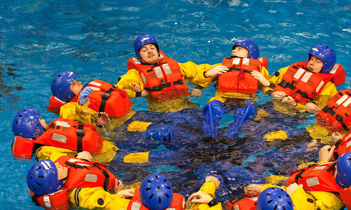 A group of people wearing life jackets and helmets form a circle while floating in a pool, resembling a strategic military formation. A person in a white helmet watches nearby.