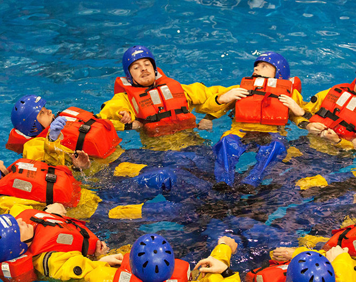 A group of people wearing life jackets and helmets form a circle while floating in a pool, resembling a strategic military formation. A person in a white helmet watches nearby.