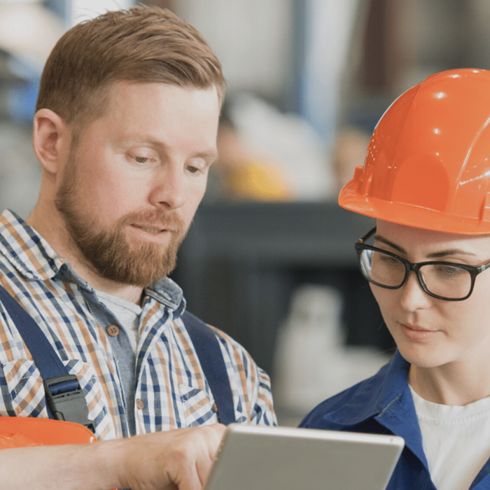 Two workers in a factory setting. One, in plaid shirt, is pointing at a tablet screen, while the other, in a blue uniform and orange hardhat, listens attentively.