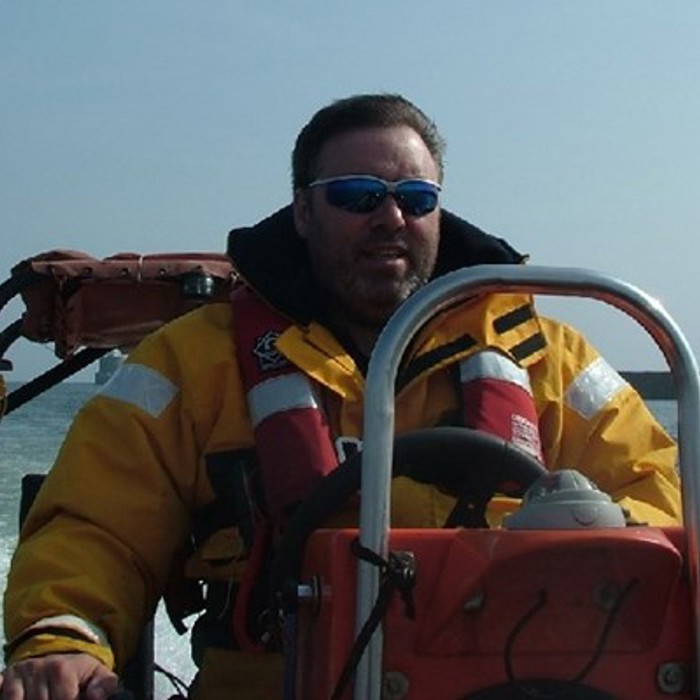 A man in a yellow jacket and blue sunglasses pilots a small boat on the water with a coastline visible in the background.