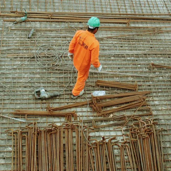 A construction worker in an orange uniform and green helmet walks over a rebar and grid mesh floor, surrounded by metal rods and cables.