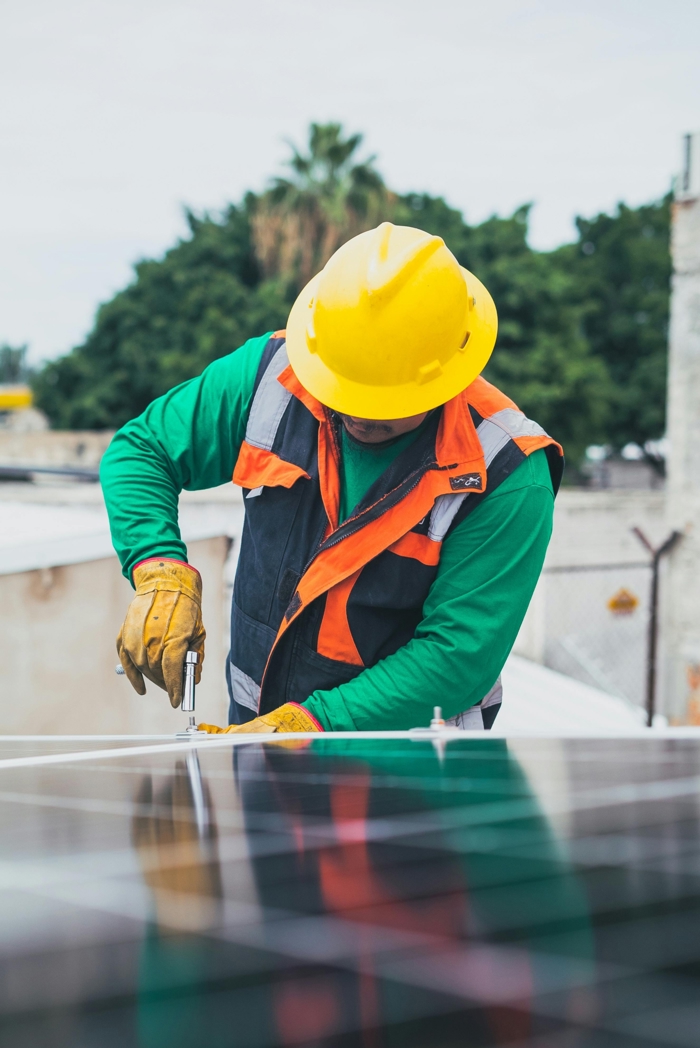 Worker in a yellow hard hat and safety vest installing solar panels outdoors.