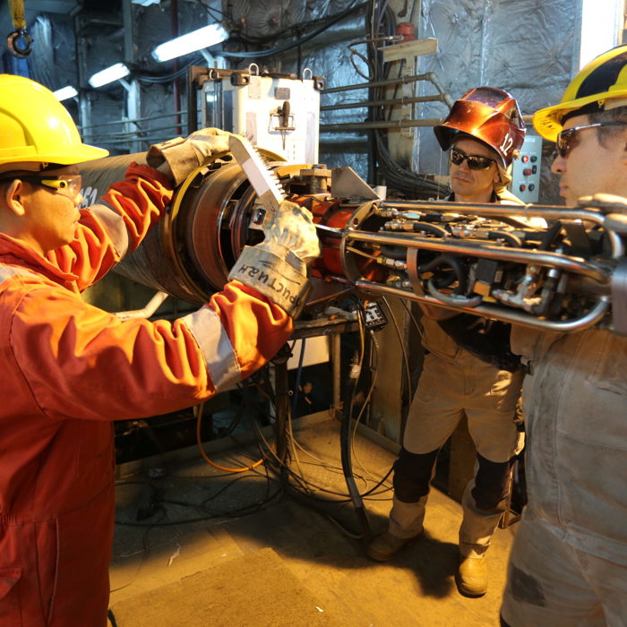 Three workers in protective gear are participating in training, collaborating in an industrial setting while handling machinery parts with tools and equipment around.