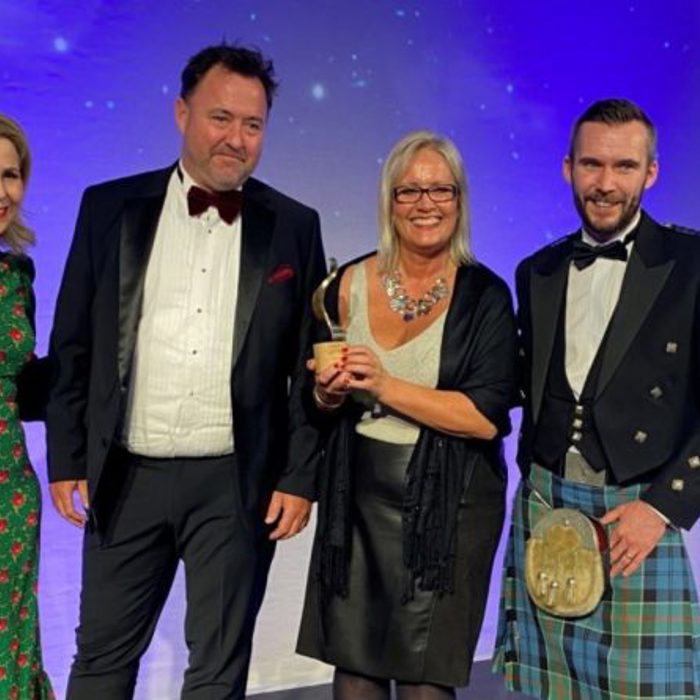 Four people at an event, with one woman in the center holding a trophy. Three individuals are wearing formal attire, including tuxedos and a kilt, while the woman on the left wears a green dress with red accents.