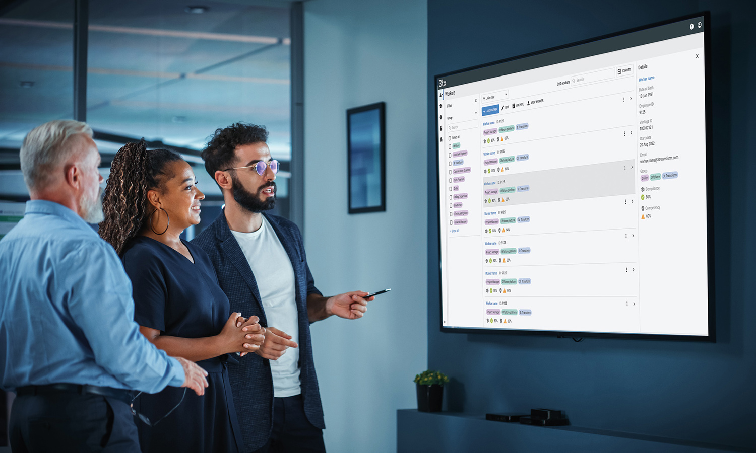Three colleagues stand in front of a large screen displaying project management software, discussing the details and pointing at the screen.