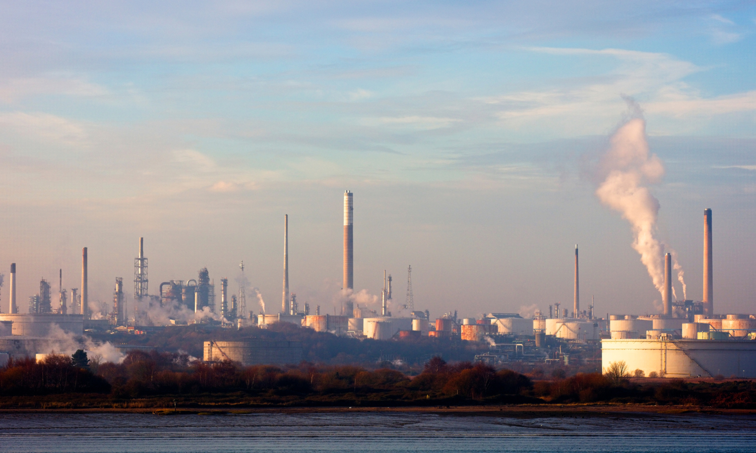 An industrial refinery landscape sprawls beneath a blue sky with scattered clouds, its chimneys puffing smoke like sentinels in training, synchronized and steadfast.