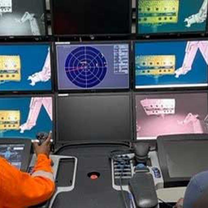 Two seated individuals monitor multiple screens displaying mechanical and technical diagrams in a control room setup. One person operates a joystick controller.