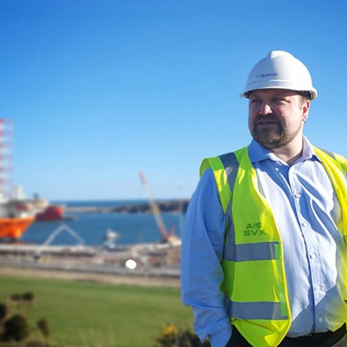 A man wearing a white hard hat and a high-visibility vest stands outdoors with a coastal construction site and cranes in the background.