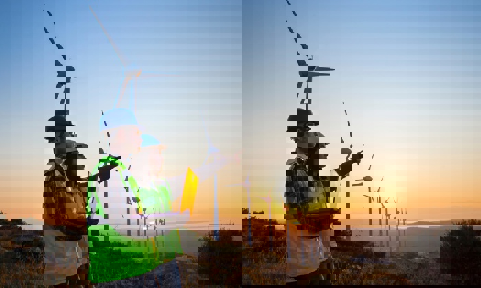 Two workers in safety gear stand with a laptop, pointing towards wind turbines at sunset on a hillside.