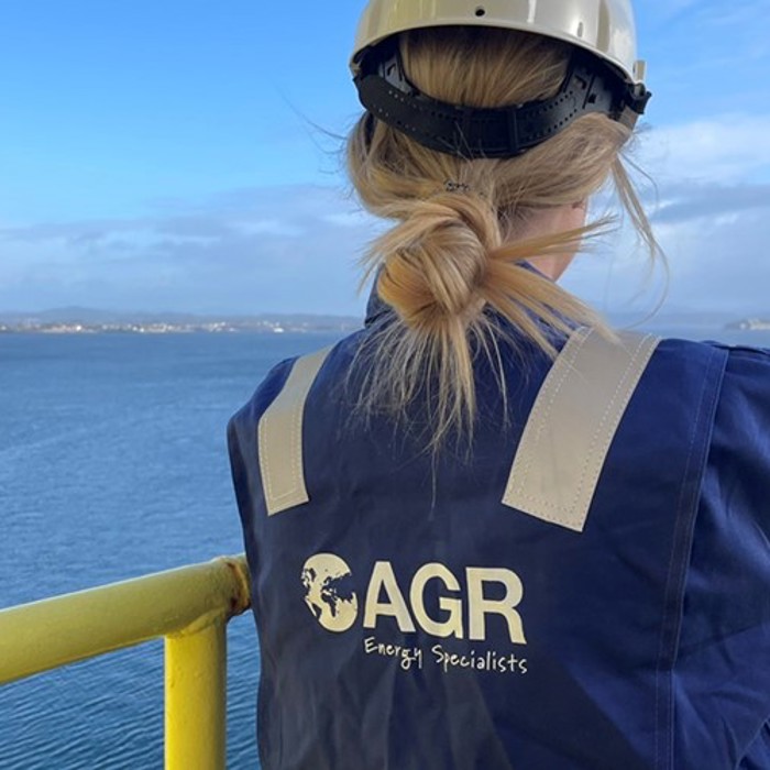 A person wearing a hard hat and a blue jacket with the logo "AGR Energy Specialists" stands on a yellow railing, overlooking a body of water.
