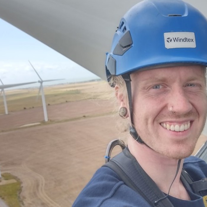 A person wearing a blue helmet labeled "Windtex" takes a selfie from a high vantage point with wind turbines and wide fields visible in the background.