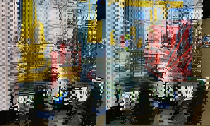 Control room setup with multiple screens and workstations displaying a detailed drilling rig simulation. Operators' chairs and monitors are in the foreground, while the virtual rig scene is in the background.