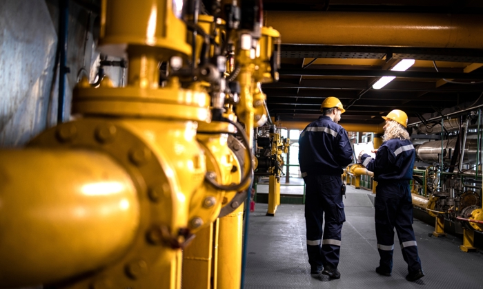 Two engineers in safety gear and helmets analyze documents amid yellow industrial pipes and machinery in a factory setting.